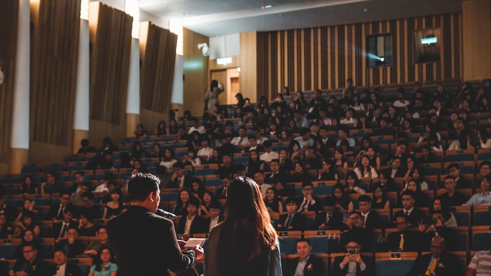 A speaker facing a large seated audience in an auditorium